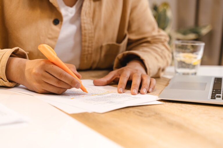 Person highlighting notes at a desk with a laptop and glass of water nearby.