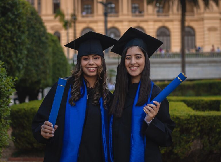 Two young women in graduation gowns smiling and holding diplomas outdoors.