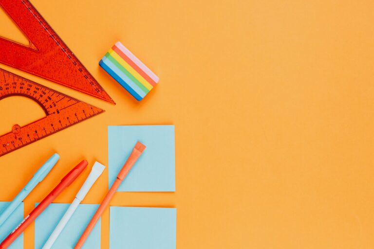 Top view of vibrant stationery essentials including rulers, pens, and sticky notes on an orange background.